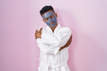 Young hispanic man wearing beauty face mask and bath robe hugging oneself happy and positive, smiling confident. self love and self care