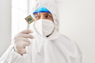Young latin man scientist wearing covid protection uniform holding cpu processor chip at laboratory