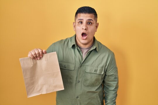 Hispanic young man holding take away paper bag scared and amazed with open mouth for surprise, disbelief face
