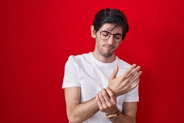 Young hispanic man standing over red background suffering pain on hands and fingers, arthritis...