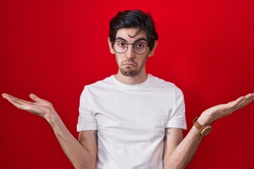 Young hispanic man standing over red background clueless and confused expression with arms and...
