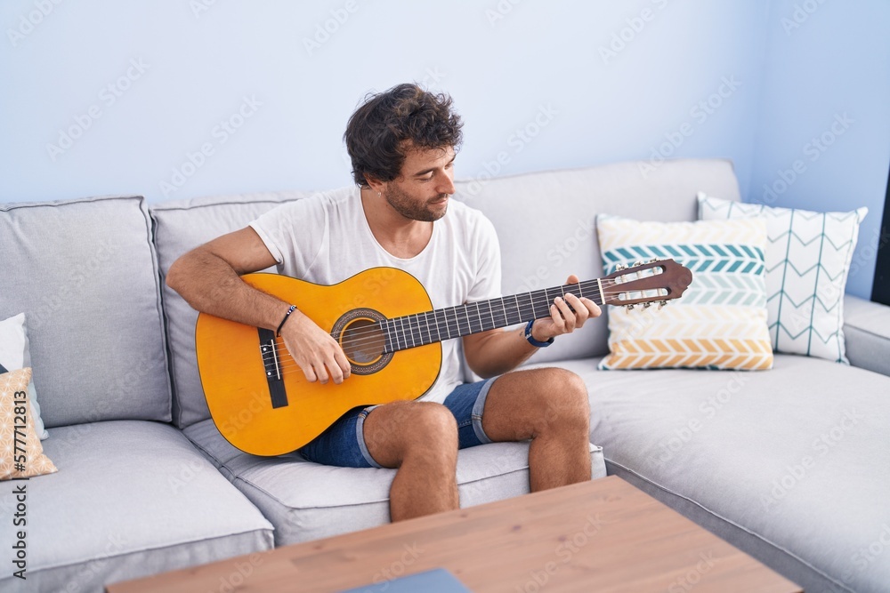 Poster Young hispanic man playing classical guitar sitting on sofa at home