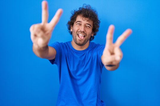 Hispanic young man standing over blue background smiling with tongue out showing fingers of both hands doing victory sign. number two.