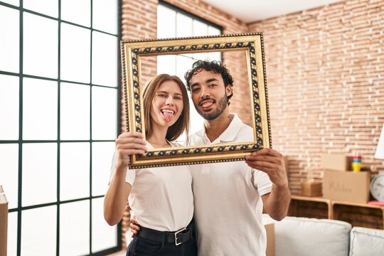 Young two people holding empty frame together sticking tongue out happy with funny expression.