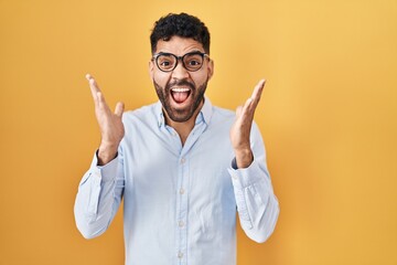 Hispanic man with beard standing over yellow background celebrating crazy and amazed for success with arms raised and open eyes screaming excited. winner concept