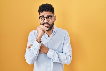 Hispanic man with beard standing over yellow background looking stressed and nervous with hands on...