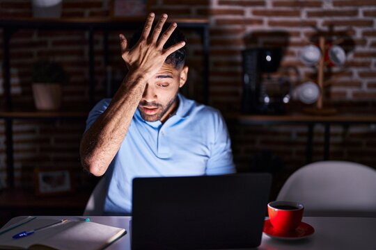 Hispanic man with beard using laptop at night surprised with hand on head for mistake, remember error. forgot, bad memory concept.