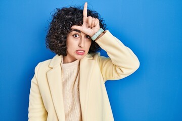 Young brunette woman with curly hair standing over blue background making fun of people with...