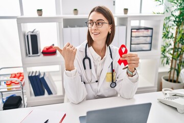 Young caucasian doctor woman holding support red ribbon pointing thumb up to the side smiling happy with open mouth