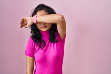 Young asian woman standing over pink background covering eyes with arm, looking serious and sad. sightless, hiding and rejection concept