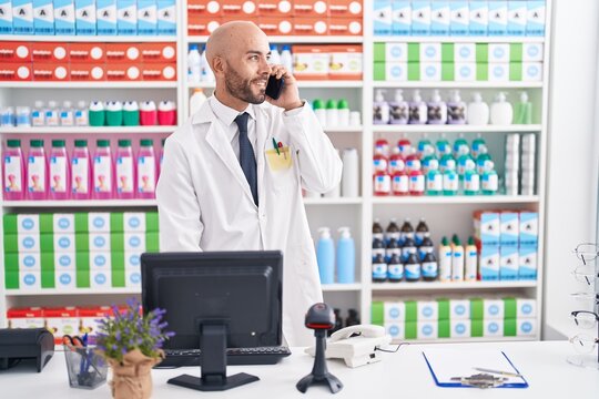 Young bald man pharmacist talking on smartphone using computer at pharmacy