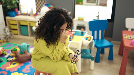 Middle age hispanic woman preschool teacher stressed sitting on chair at kindergarten
