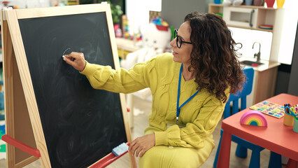 Middle age hispanic woman preschool teacher writing on blackboard at kindergarten