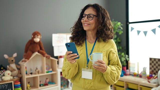 Middle age hispanic woman preschool teacher using smartphone drinking coffee at kindergarten