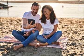 Fototapeta na wymiar Middle age man and woman couple using touchpad sitting on sand at seaside