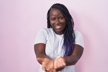 Young african woman standing over pink background smiling with hands palms together receiving or...