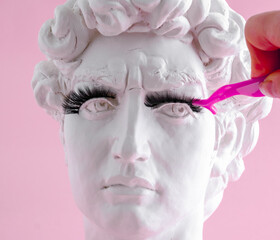 Plaster statue head wearing eyelashes pink background. Beauty procedure concept.