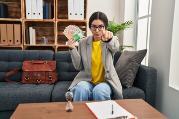 Young hispanic woman working at consultation office holding money pointing with finger to the camera and to you, confident gesture looking serious