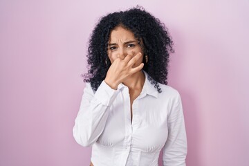 Fototapeta na wymiar Hispanic woman with curly hair standing over pink background smelling something stinky and disgusting, intolerable smell, holding breath with fingers on nose. bad smell