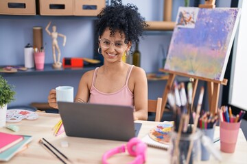 Young hispanic woman artist using laptop drinking coffee at art studio