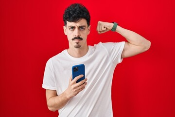 Hispanic man using smartphone over red background strong person showing arm muscle, confident and proud of power