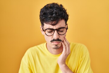 Hispanic man wearing glasses standing over yellow background touching mouth with hand with painful expression because of toothache or dental illness on teeth. dentist