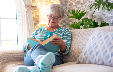 Portrait of beautiful smiling senior woman sitting on sofa at home reading a book - concept of...