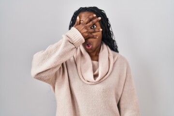 African woman standing over white background peeking in shock covering face and eyes with hand,...