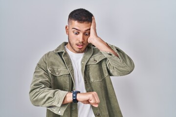 Young hispanic man standing over isolated background looking at the watch time worried, afraid of getting late