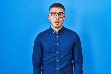 Obraz premium Young hispanic man wearing glasses over blue background afraid and shocked with surprise expression, fear and excited face.