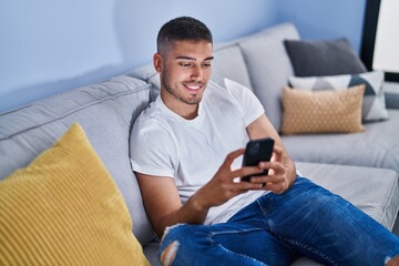 Young hispanic man using smartphone sitting on sofa at home
