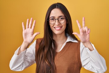 Young brunette woman standing over yellow background wearing glasses showing and pointing up with fingers number eight while smiling confident and happy.