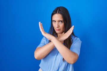 Young brunette woman standing over blue background rejection expression crossing arms doing negative sign, angry face