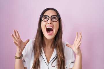Young brunette woman wearing glasses standing over pink background crazy and mad shouting and...