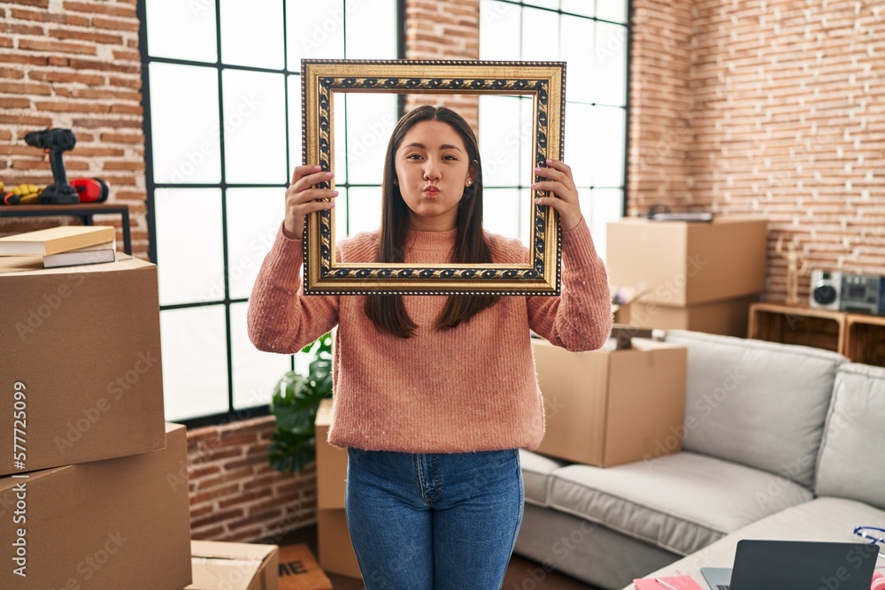 Sticker young latin woman moving to a new home holding empty frame puffing cheeks with funny face. mouth inf