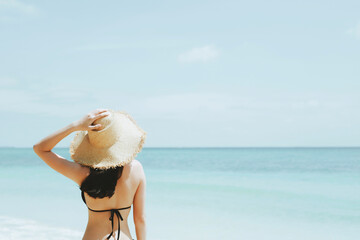 Fototapeta na wymiar backside young woman skin tan in sunhat and bikini standing with her arms raised to her head enjoying on the beach vacation travel. and enjoy life at sea looking view of beach ocean on hot summer day.