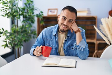 Young hispanic man drinking coffee reading book at home