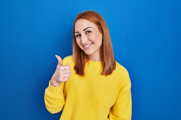 Young woman standing over blue background doing happy thumbs up gesture with hand. approving expression looking at the camera showing success.