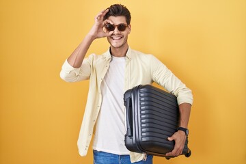 Young hispanic man holding suitcase going on summer vacation doing ok gesture with hand smiling,...