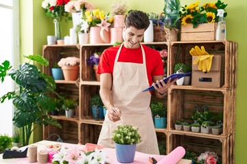 Young hispanic man florist using touchpad touching plant of shelving at flower shop