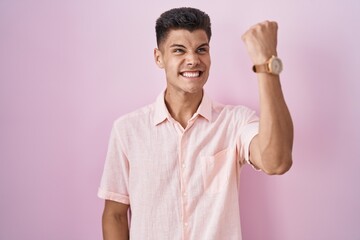 Young hispanic man standing over pink background angry and mad raising fist frustrated and furious while shouting with anger. rage and aggressive concept.