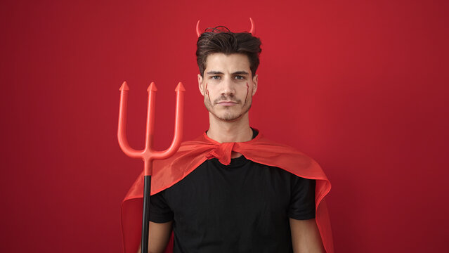 Young hispanic man wearing devil costume holding trident over isolated red background