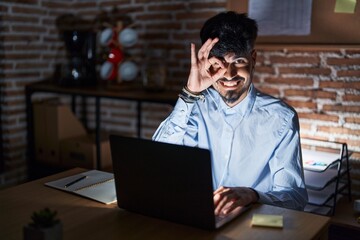 Young hispanic man with beard working at the office at night doing ok gesture with hand smiling, eye looking through fingers with happy face.