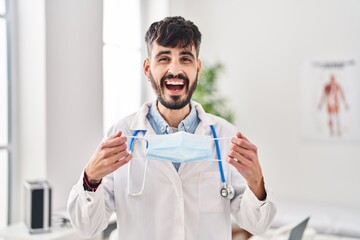 Young hispanic doctor man with beard holding safety mask smiling and laughing hard out loud because funny crazy joke.
