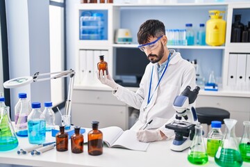 Young hispanic man scientist writing on notebook holding bottle at laboratory