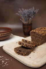 Black Bread with cereals Sliced on a Wooden cutting Close up Board against a brown background. Next to the bread is a Clay Plate, fork, spoon and white cotton tablecloth