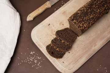 A Top View of Black Bread with cereals Sliced on a wooden cutting board against a brown background. A Knife and a white cotton tablecloth Lie next to the Bread