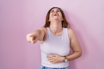 Brunette woman standing over pink background laughing at you, pointing finger to the camera with...