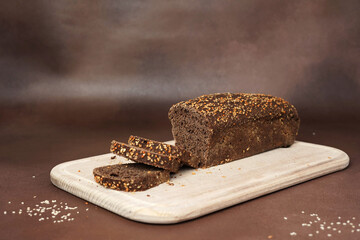Black Bread with Cereals sliced on a wooden cutting board against a Brown Background. Preparing the Dinner Table