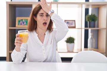 Brunette woman drinking glass of orange juice surprised with hand on head for mistake, remember error. forgot, bad memory concept.